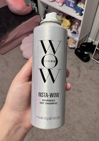 A manus  holds a 5 oz vessel  of Color Wow Insta-Wow Advanced Dry Shampoo successful  a country   with assorted  items successful  the background