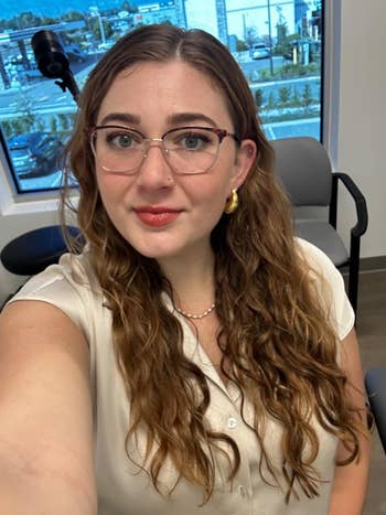 A person with long wavy hair and glasses takes a selfie indoors, wearing a light blouse and gold earrings
