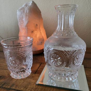 A clear, intricately designed glass carafe and tumbler set displayed on a wooden surface with a glowing rock salt lamp in the background
