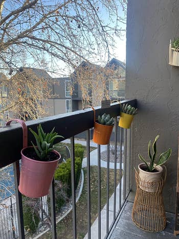 Balcony railing with hanging planters, adjacent to a potted plant on a stand, overlooking residential area. Ideal for small-space gardening