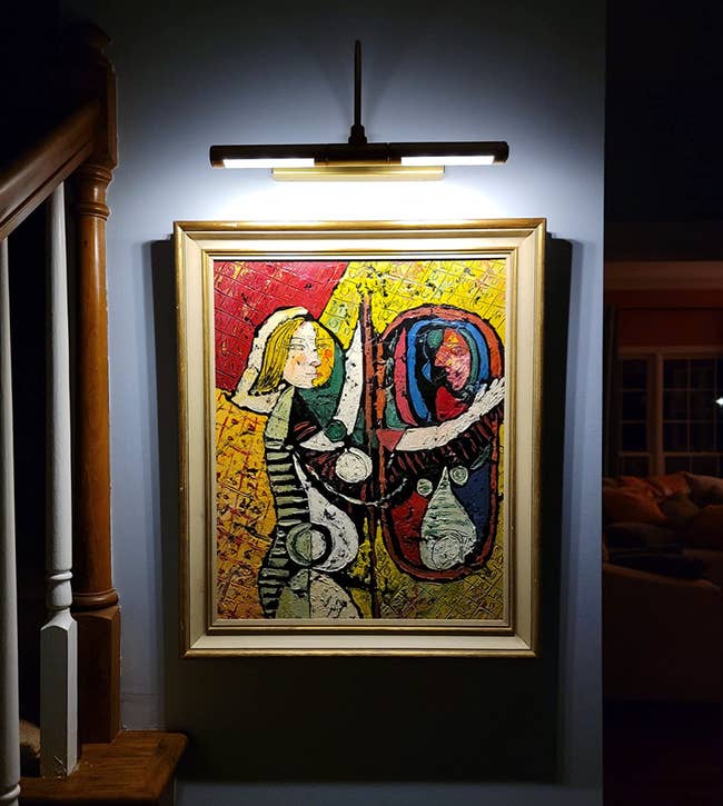 Painting with abstract figures displayed under a light in a dark room, hung on a wall near a stair banister