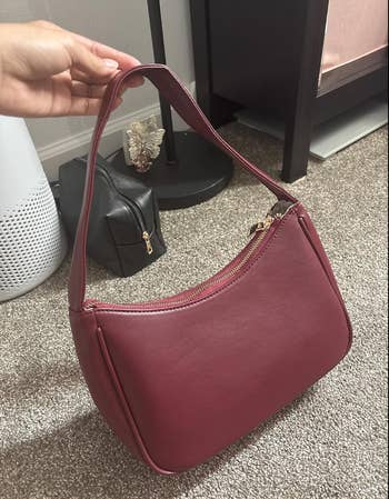 Hand holding a medium-sized handbag with zip closure, placed adjacent  to a lamp and achromatic  purse, suitable for shopping