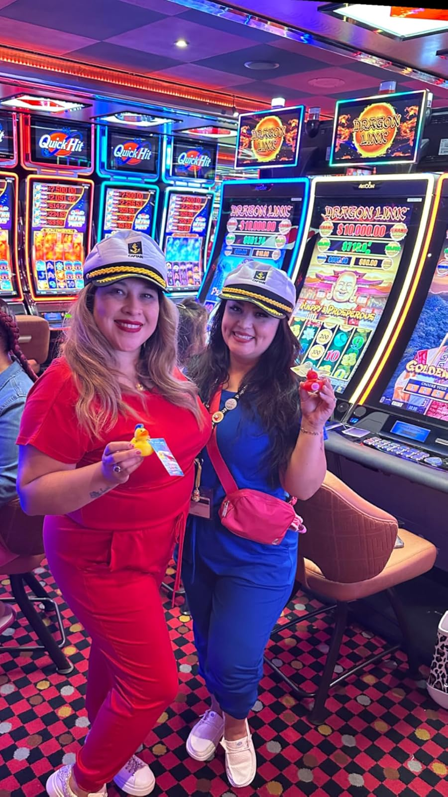 Two people in sailor hats pose holding casino items in front of slot machines, dressed in casual red and blue outfits with sneakers