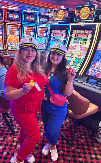 Two people in sailor hats pose holding casino items in front of slot machines, dressed in casual red and blue outfits with sneakers