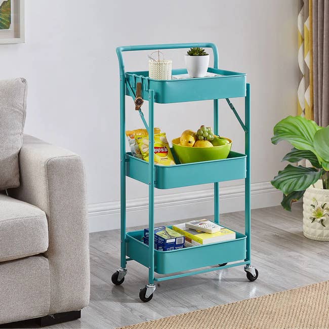 Rolling utility cart with three shelves holds snacks, fruit, books, and decor. Near sofa and plants, suitable for home storage or kitchen use
