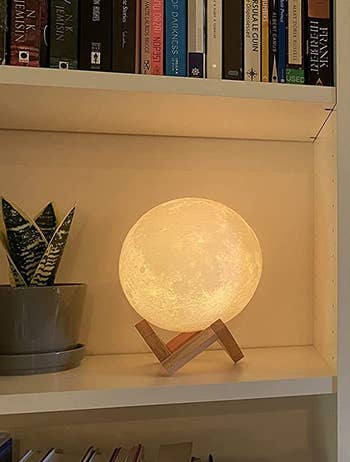 reviewer photo of the lit moon lamp on its wooden stand on a bookshelf