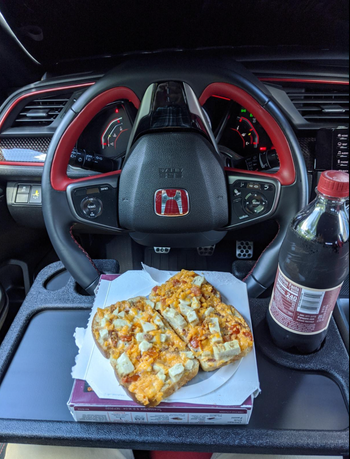 A black plastlc floating tray attached to a steering wheel with a sandwich and coke on it