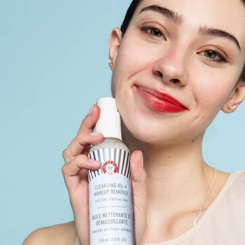model with half face makeup removed, holding cleanser
