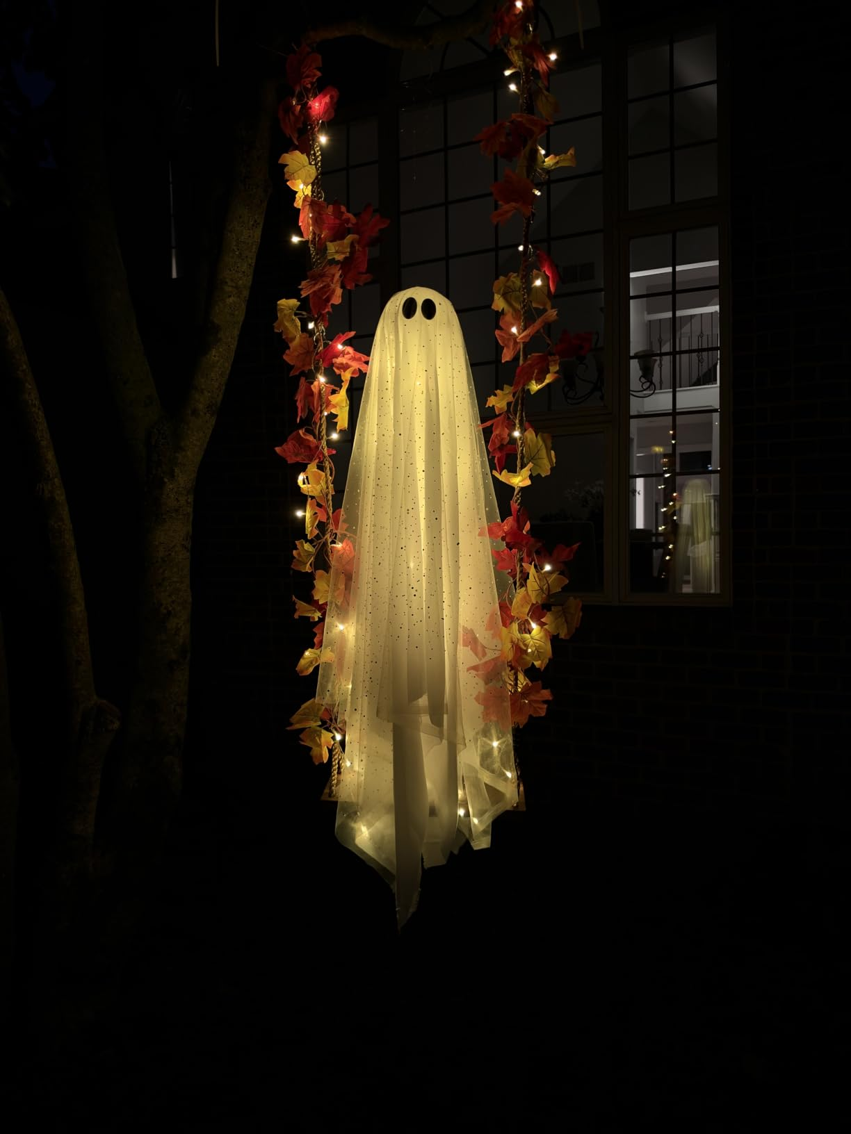 Ghost decoration with lights hanging from a tree, surrounded by autumn leaves, creating a spooky ambiance for Halloween shopping