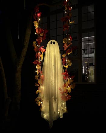 Ghost decoration with lights hanging from a tree, surrounded by autumn leaves, creating a spooky ambiance for Halloween shopping