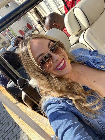 Woman in sunglasses and denim jacket smiles for a selfie in a convertible