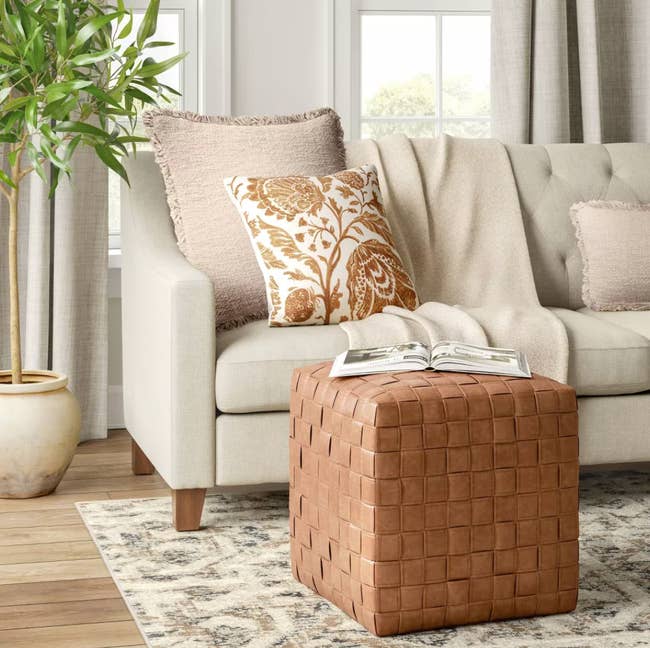 A woven leather ottoman in a living room, paired with a beige sofa with a floral cushion