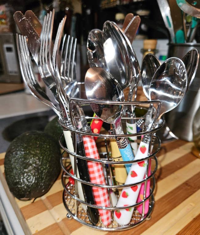 Assorted cutlery organized in a metal countertop holder next to an avocado, suggesting kitchenware organization for shopping