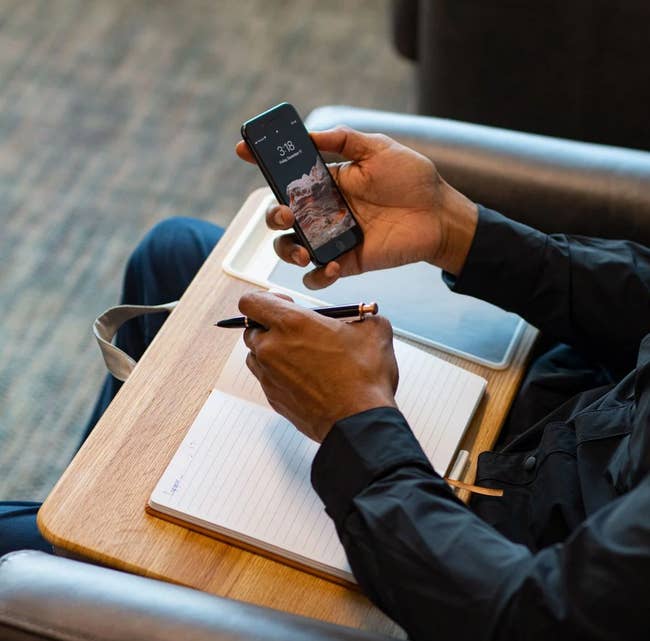Person holding a smartphone and penning successful a notebook, seated astatine a table, suggesting multitasking oregon readying portion buying online