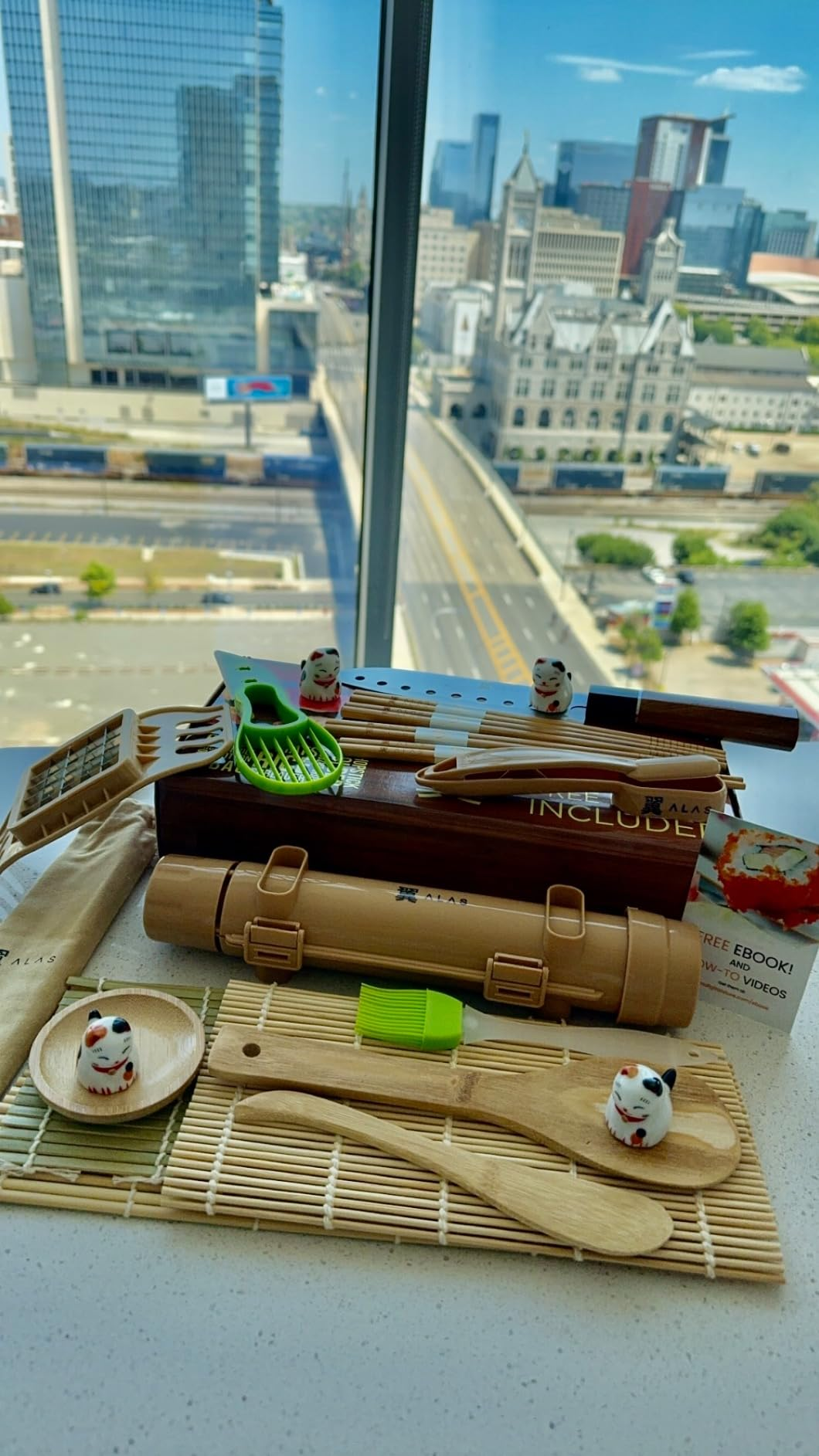 Bamboo sushi kit with utensils displayed on a table, overlooking a cityscape through a window in the background