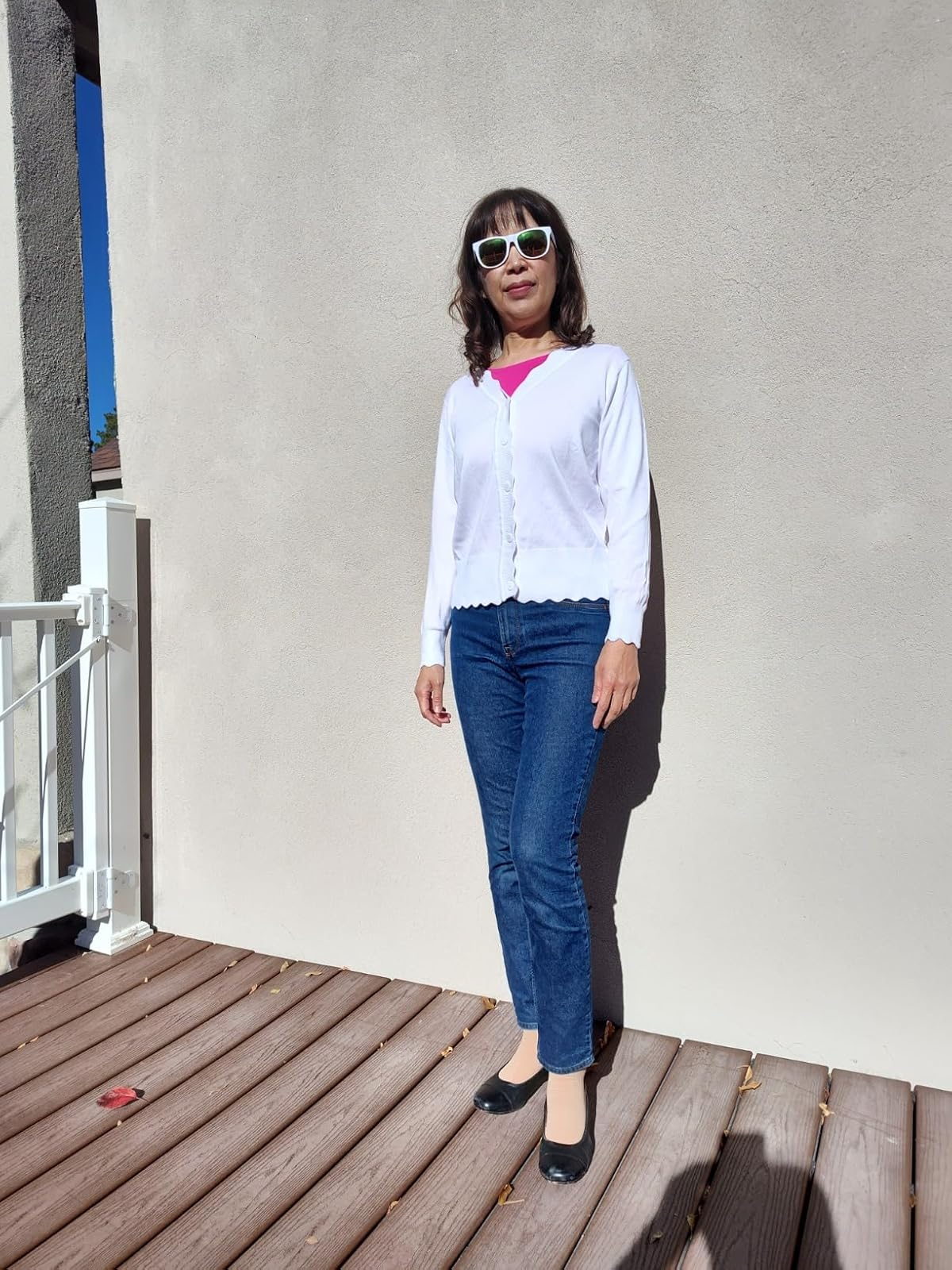 A woman in sunglasses stands on a deck wearing a white cardigan, pink top, blue jeans, and black shoes, showcasing casual style
