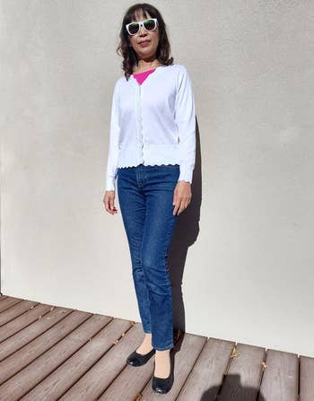 A woman in sunglasses stands on a deck wearing a white cardigan, pink top, blue jeans, and black shoes, showcasing casual style