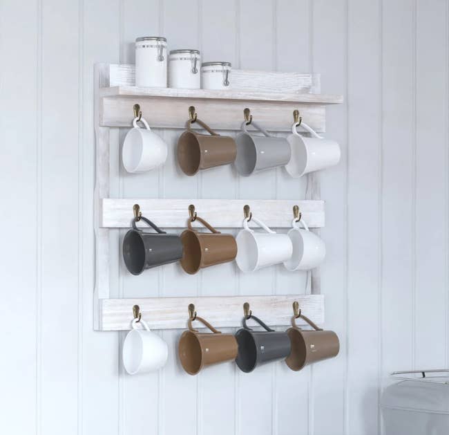 White rustic wood hanging mug organizer holding twelve mugs (four on each of the three rows) with a top shelf holding sugar containers 