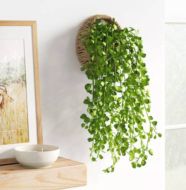 A woven wall planter with fake vines falling out attached to a white wall next to a wooden floating shelf
