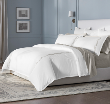White bedding with gray scalloped vines embroidered on the duvet and white matching pillows on a white bed frame