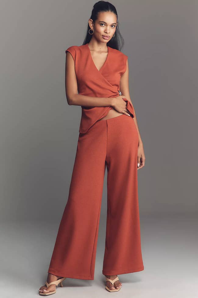 Person wearing a stylish, draped orangish ensemble with wide-leg pants and open-toed sandals, posing confidently