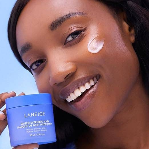 A person smiling with a small amount of Laneige Water Sleeping Mask applied to their cheek, holding the product container next to their face