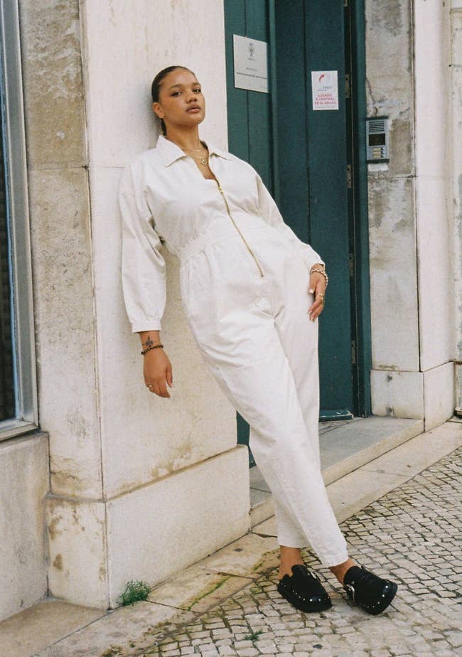 Person in a white jumpsuit and black studded loafers posing against a wall