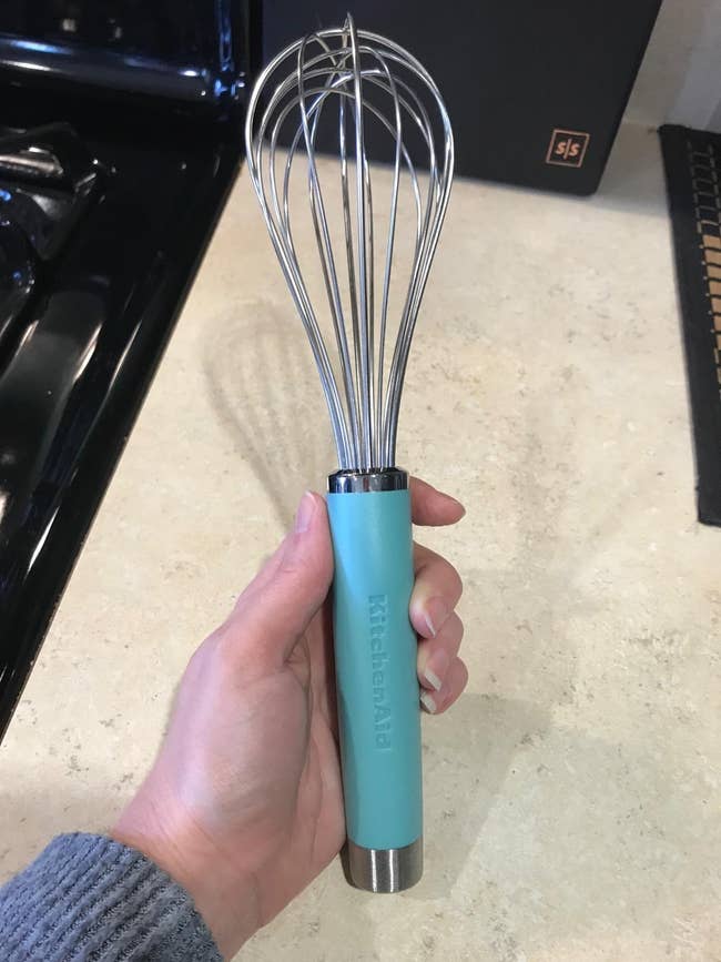 Person holding a KitchenAid whisk with a stainless steel finish and aqua handle