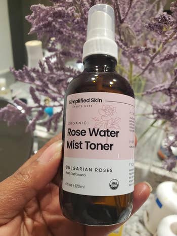 reviewer holding a vessel  of Simplified Skin Rose Water Mist Toner, surrounded by lavender flowers