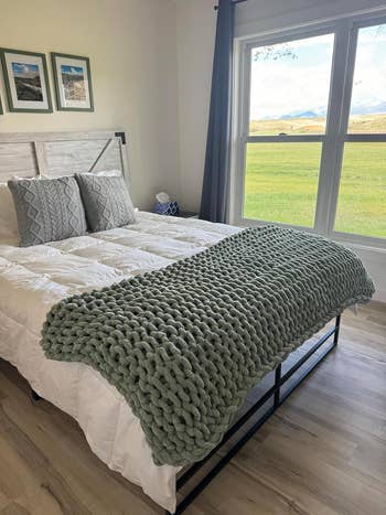 Cozy bedroom featuring a modern bed with a chunky knit blanket and decorative pillows, next to a large window with a serene outdoor view