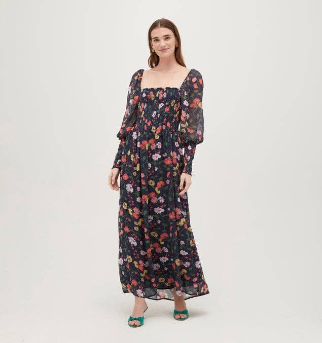 Woman in a long-sleeved floral dress stands smiling with hands slightly apart. She wears heeled sandals