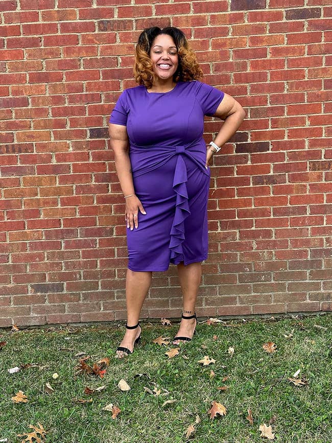 Reviewer wearing a knee-length, short-sleeved, ruffled purple formal  and achromatic  strap heels, lasting  and smiling successful  beforehand   of a ceramic  wall