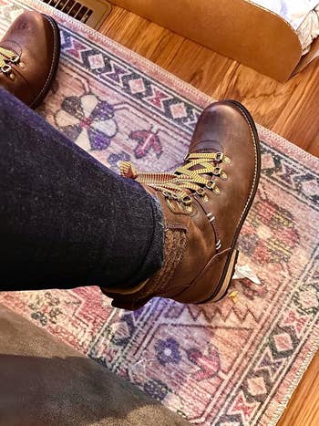 Brown hiking boots with laces and rugged soles worn with acheronian  jeans, shown indoors connected  a patterned rug