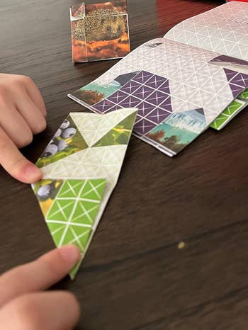 Hands folding patterned origami insubstantial  connected  a table, surrounded by assorted  designs