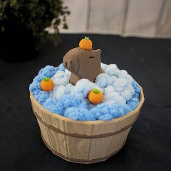 Capybara-shaped toy in a wooden bucket, surrounded by curled up fabric coasters to look like foaming water 