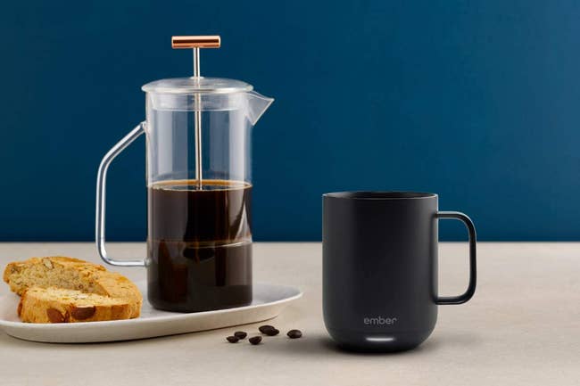 The black mug on a table next to a french press and biscotti