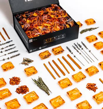 An unfastened  container  with amber-colored fragrance pods, arranged with cinnamon sticks, lavender, and herbs, suggesting a luxurious scent collection