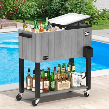 Outdoor rolling cooler by a pool, filled with assorted bottled drinks and a tray beneath with much bottles, suitable for poolside gatherings