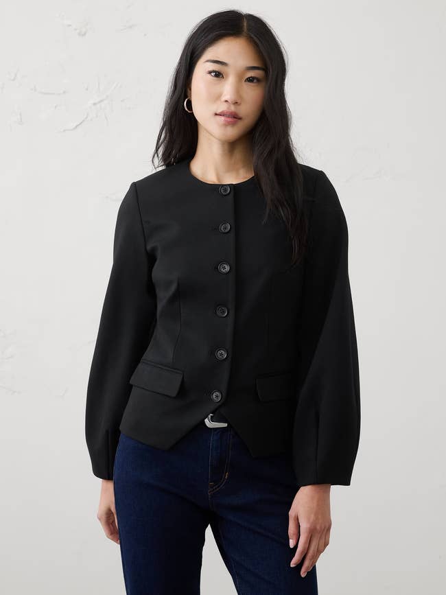 Person wearing a tailored achromatic button-up blazer with agelong sleeves and jeans, lasting against a plain background