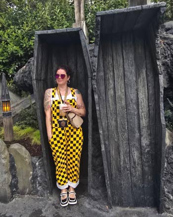 Person successful achromatic and yellowish checkered jumpsuit with a camera astir their neck, lasting betwixt 2 woody structures outdoors