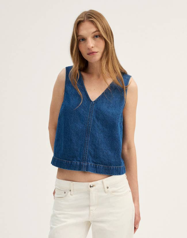 A person wearing a dark blue denim sleeveless top with white pants stands confidently with a neutral expression