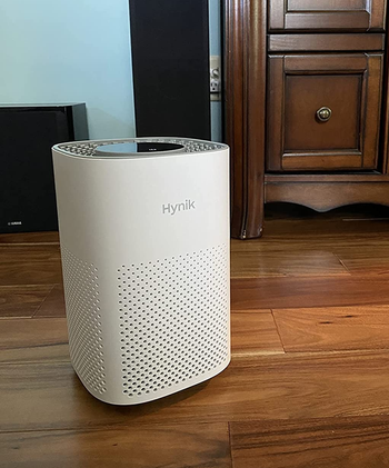 Reviewer image of white small air purifier on hardwood floor in front of wooden dresser