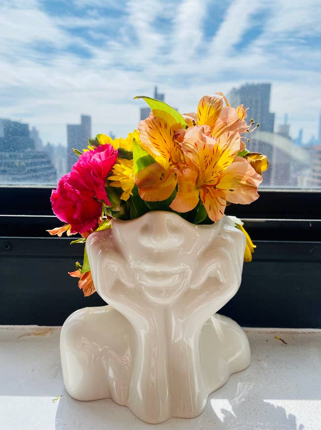 Vase with floral arrangement on a windowsill, shaped like a human head, with city skyline in background. Perfect for modern home decor