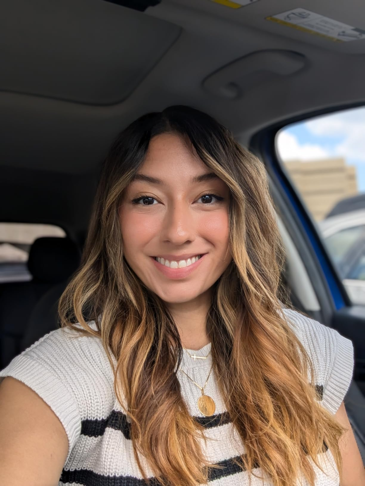 Reviewer successful  a car   smiling, wearing a knitted apical  with achromatic  stripes and layered necklaces, looking casual and relaxed