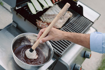 model's hand holding the basting mop, which is in a bowl of sauce, to show how long the handle is
