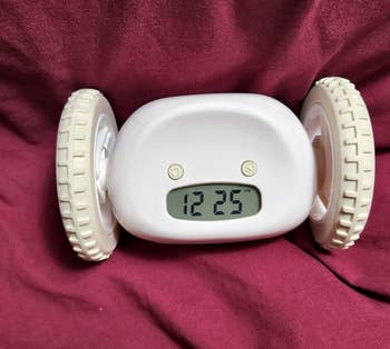 White rolling alarm clock with digital display showing 12:25, set against a draped fabric background