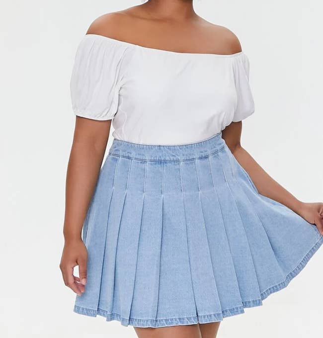 Model is wearing a white top with a light denim pleated mini skirt