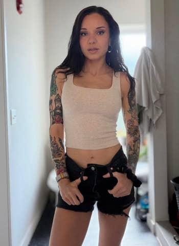 Person with tattoos successful  achromatic  vessel  apical  and achromatic  shorts stands confidently successful  room, hands connected  hips. Casual, stylish manner  featured successful  buying  context