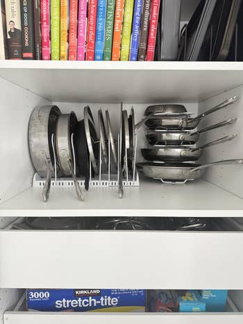 Neatly organized kitchen cabinet with stacked frying pans, cookbooks, and a roll of stretch wrap on the bottom shelf