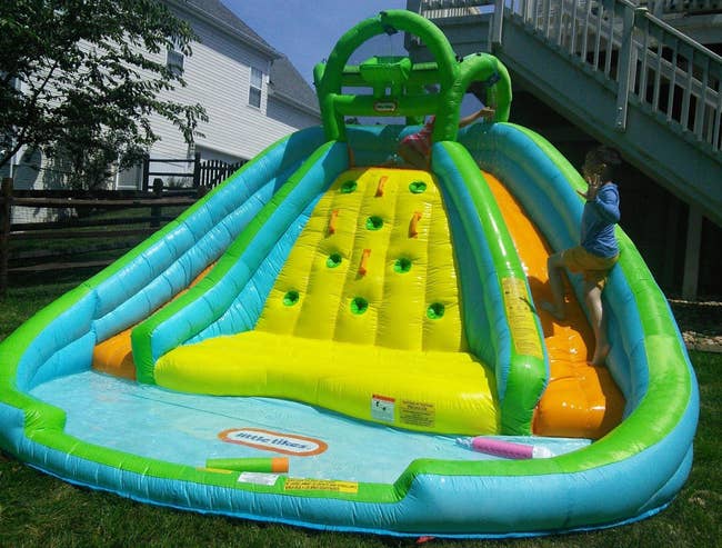 Children climbing a colorful inflatable h2o descent with climbing grips successful a backyard setting. A excavation of h2o is astatine the base